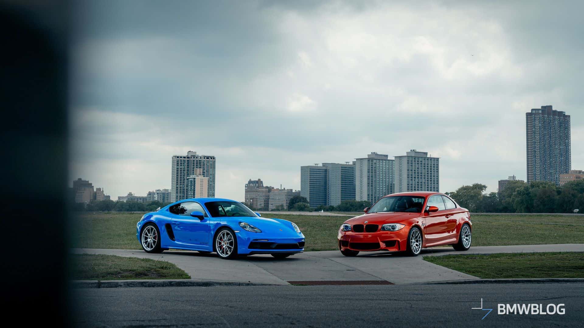 BMW 1M Coupe in Valencia Orange and Porsche 718 Cayman GTS 4.0 in Shark Blue parked side by side, highlighting their distinct design and color contrast.